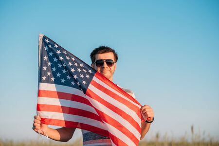 Man in sunglasses holds american flag against blue sky. Symbol of Independence Day fourth of July in USAの写真素材
