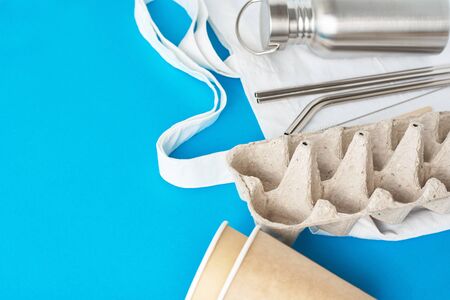 Zero waste concept. Eco friendly reusable items on natural shopping bag. Paper egg tray, coffee cups, aluminum bottle and metal tubes on blue backgroundの写真素材