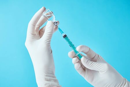 Hands in gloves hold an injection syringe and medicine vial ampoule against blue background.  Healthcare, medical and virus protection conceptの写真素材