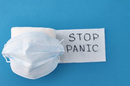 Panic buying Covid-19 Coronavirus outbreak concept. Roll of toilet paper with inscription stop panic and surgical mask on blue background.の写真素材