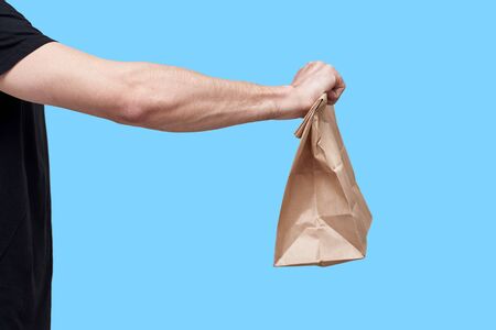 Food delivery concept. Courier in black uniform hold paper bag with take away food on blue backgroundの写真素材