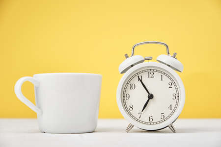 Morning time concept. White retro alarm clock and cup on yellow background