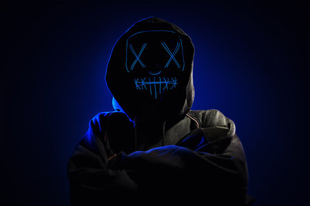 Anonymous man in a hoodie hiding face behind neon glow scary mask on dark background. Horror conceptの写真素材