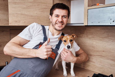 Happy worker man in uniform with screwdriver in hand smile, shows thums up gesture and hugging dog on the kitchen background. Maintenance of kitchen furnitureの写真素材