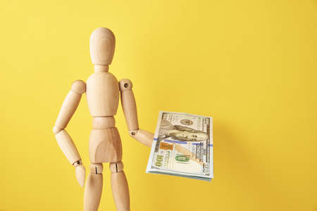 Figure of wooden human holds cash money on yellow background. Wooden doll with dollar banknotes on yellow background. Business and finance conceptの写真素材