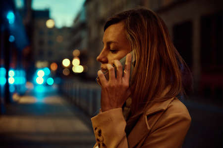 Woman in coat walks at night city street and use smartphone. City life