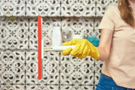 Woman in yellow rubber gloves cleans bathroom shower cabine with scraper. House cleaningの写真素材