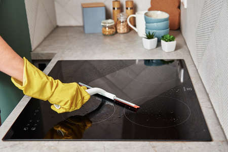 Cleaning induction stove. Woman in yellow rubber gloves cleans kitchen induction hob with cleaning sparay. Clear kitchen applianceの写真素材