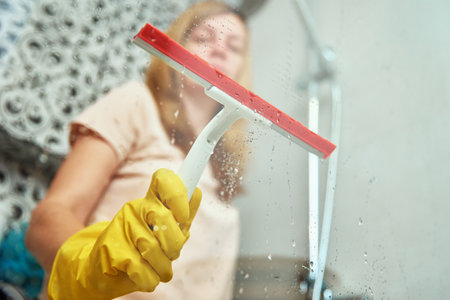 Woman in yellow rubber gloves cleans bathroom shower cabine with scraper. House cleaningの写真素材