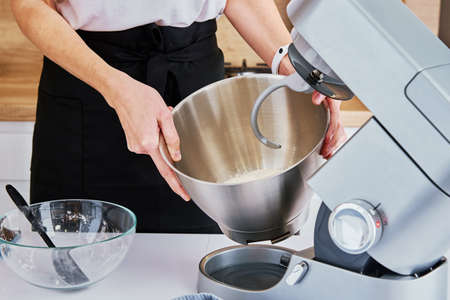 Woman prepares dough using kitchen mixer, pouring ingredients into steel bowl. Cooking homemade cake. Modern kitchen appliancesの写真素材