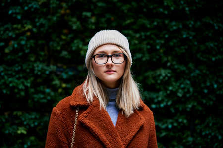 Portrait of woman in coat and eyeglasses standing near green natural backgroundの写真素材
