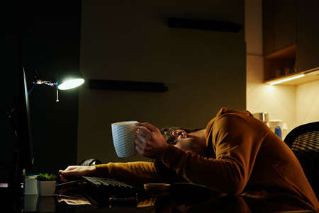 Man sleeping at home office workplace with cup of coffeee in his hand. Freelancer remotely working...