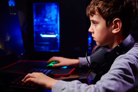 Teenager boy play computer video game in dark room, use neon colored rgb mechanical keyboard,...