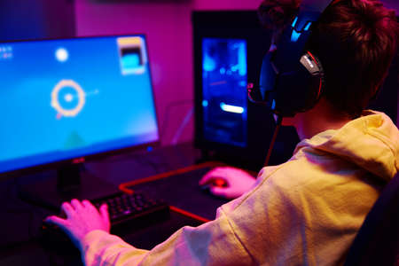 Teenager boy play computer video game in dark room, use neon colored rgb mechanical keyboard,...