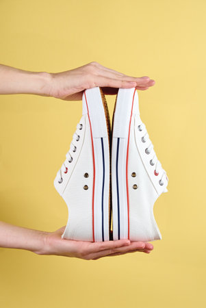 Pair of sport sneakers in woman hands against yellow background, Casual trendy shoes closeup,...