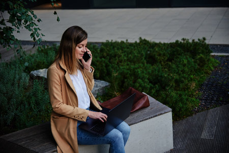 Woman remotely working, sitting on bench at city street and using laptop. Female freelancer work...