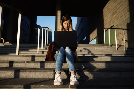 Woman sitting on steps outdoors and using laptop. Concept of remote working and online education.の写真素材