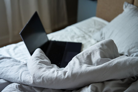 Opened laptop in unmade bed with pillows and crumpled bedsheets at morning. Comfortable workplace for freelancer, work from homeの写真素材