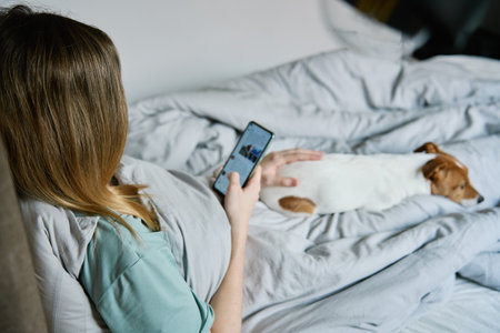 Woman with cute dog chilling in bed at morning and use smartphone, lazy morning. Spending time together, pet affectionの写真素材