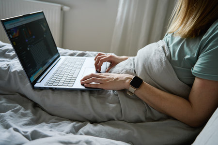 Millennial female freelancer lying in bed and using laptop. Woman work from home. Comfortable...