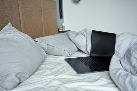 Opened laptop in unmade bed with pillows and crumpled bedsheets at morning. Comfortable workplace for freelancer, work from homeの写真素材