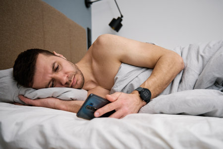 Phone addiction. Man lying in bed with smartphone. Man resting in bedroom using cellphoneの写真素材