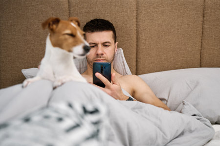 Man with cute dog chilling in bed at morning and use smartphone, lazy morning. Spending time together, pet affectionの写真素材