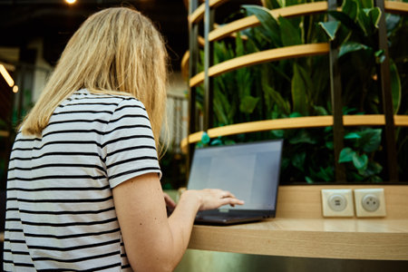 Woman using laptop in coworking with green plants. Female freelancer typing on laptop keyboard. Online work in cafeの写真素材