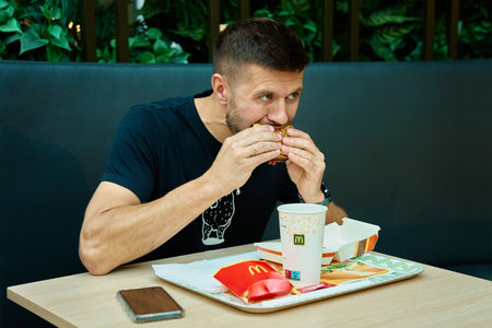 Man has breakfast in Mcdonalds restaurant. Male eating eating hamburger and french fries in Mccafe. Fast food nutrition. Wroclaw, Poland - July 2, 2023のeditorial素材