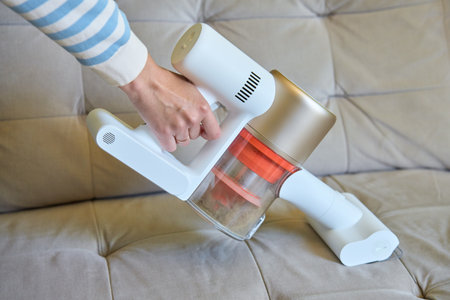 Woman is using portable cordless vacuum cleaner to clean surface of sofa in living room. House...