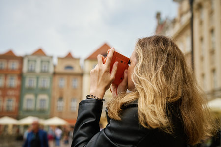 Woman taking picture using orange instant camera. Tourist captures memories during travel with vintage camera. Female walking around city streetの写真素材