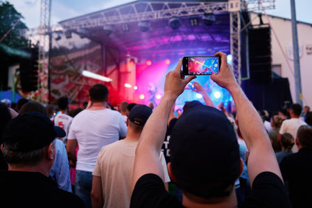 Person standing in front of stage, recording live music concert on smartphone. People crowd near...