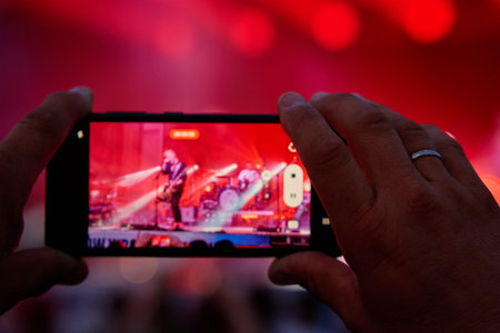 Person standing in front of stage, recording live music concert on smartphone. People crowd near stage enjoys musician band's performanceの写真素材
