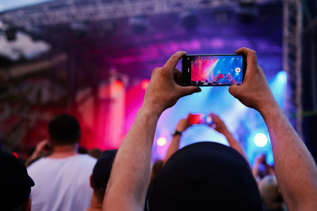 Person standing in front of stage, recording live music concert on smartphone. People crowd near...