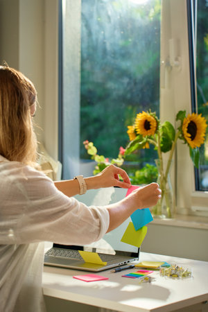 Woman is using sticky notes on her laptop screen to organize her work while working remotely from...