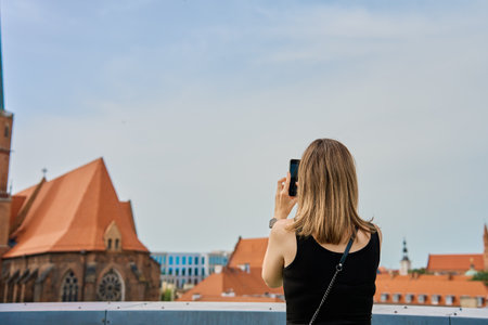 Woman taking photos of historical buildings in European city. Female tourist captures memories during travel. Woman using smartphone at city streetの写真素材