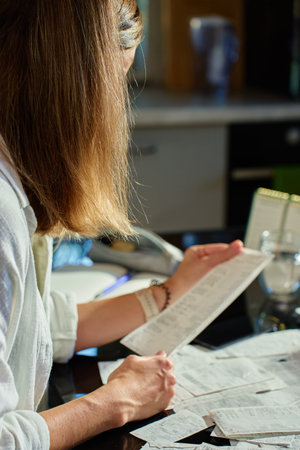 Stressed woman reviewing bills at home. Female organizing receipts, sitting at kitchen table with laptop and paper documents. Concept of budgeting and managing personal financesの写真素材