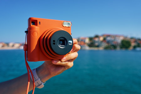 Close-up of tourist hand holding an orange instant camera in retro style with coastal town and blue sea in background. Summer travel concept. Equipment for capturing momentsの写真素材