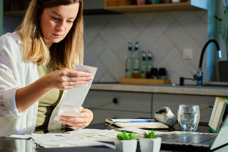 Portrait of stressed woman sitting in kitchen with laptop and stack of paper invoice. Female planning home budget, looking at finance bill and calculating expensesの写真素材