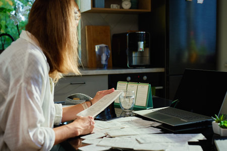 Woman sitting at kitchen table overwhelmed with bills and receipts. Financial stress and personal finance management challenges at homeの写真素材