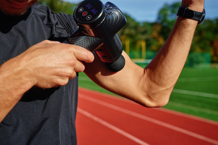 Close-up of male athlete using handheld massage gun on arm muscles for recovery after training session on stadium track. Concept of muscle relaxation and sports recoveryの写真素材
