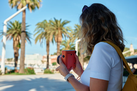 Female traveler in casual clothing taking picture in coastal town. Woman using orange retro instant camera to take photo during vacation. Tourist captures memories during travelの写真素材