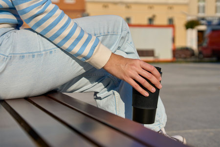 Woman in striped sweater and blue jeans enjoying her coffee break outdoors, holding black reusable coffee cupの写真素材