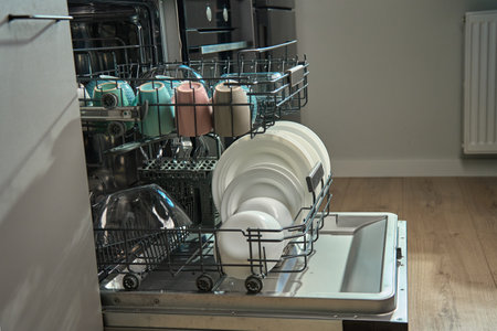 Open dishwasher in kitchen loaded clean crockery items, including white plates, bowls and cups ready...