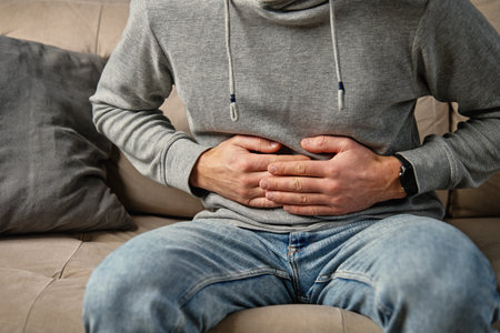 Close-up of man sitting on a couch at home, clutching his stomach, suffering from abdominal pain or cramps. Person suffering from health issues, digestion problems or food poisoningの写真素材