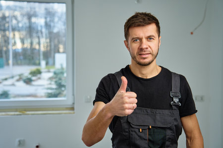 Confident construction worker in black overalls giving thumbs up in partially renovated room with large window. Concept of home improvement and renovationの写真素材