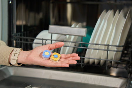 Close-up of female hand holding two dishwasher detergent tablets near open dishwasher with clean dishes inside. Essential cleaning product for effective dishwashing and hygieneの写真素材