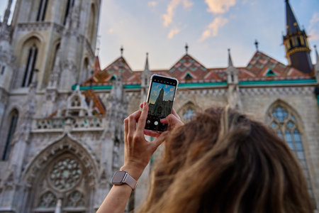 Female tourist capturing photo of historic cathedral in Budapest using smartphone. Traveler walking at city streetの写真素材