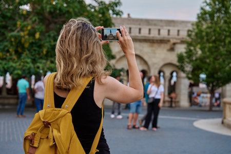 Female tourist with yellow backpack taking photo of historic landmark in Budapest during travel. Woman using smartphone to capture travel and sightseen moments in European cityの写真素材