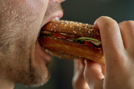 Close up of mouth takes big bite of seeded bagel filled with egg, cheese, tomato and greens. Hungry man eats sandwichの写真素材